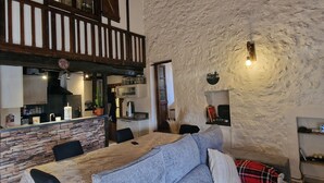 Un intérieur rustique avec un mur en pierre, des poutres en bois, un coin repas avec une table et des chaises, et une cuisine équipée d’un porte-ustensiles suspendu.