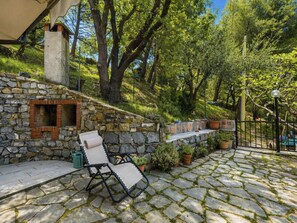 A stone patio with a brick fireplace, outdoor seating, and potted plants. - Quaranta by Interhome (Cervo)