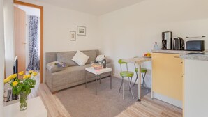 Ett kompakt vardagsrum med en soffa, ett litet matbord, en köksdel med en kaffebryggare och en vas med blommor.