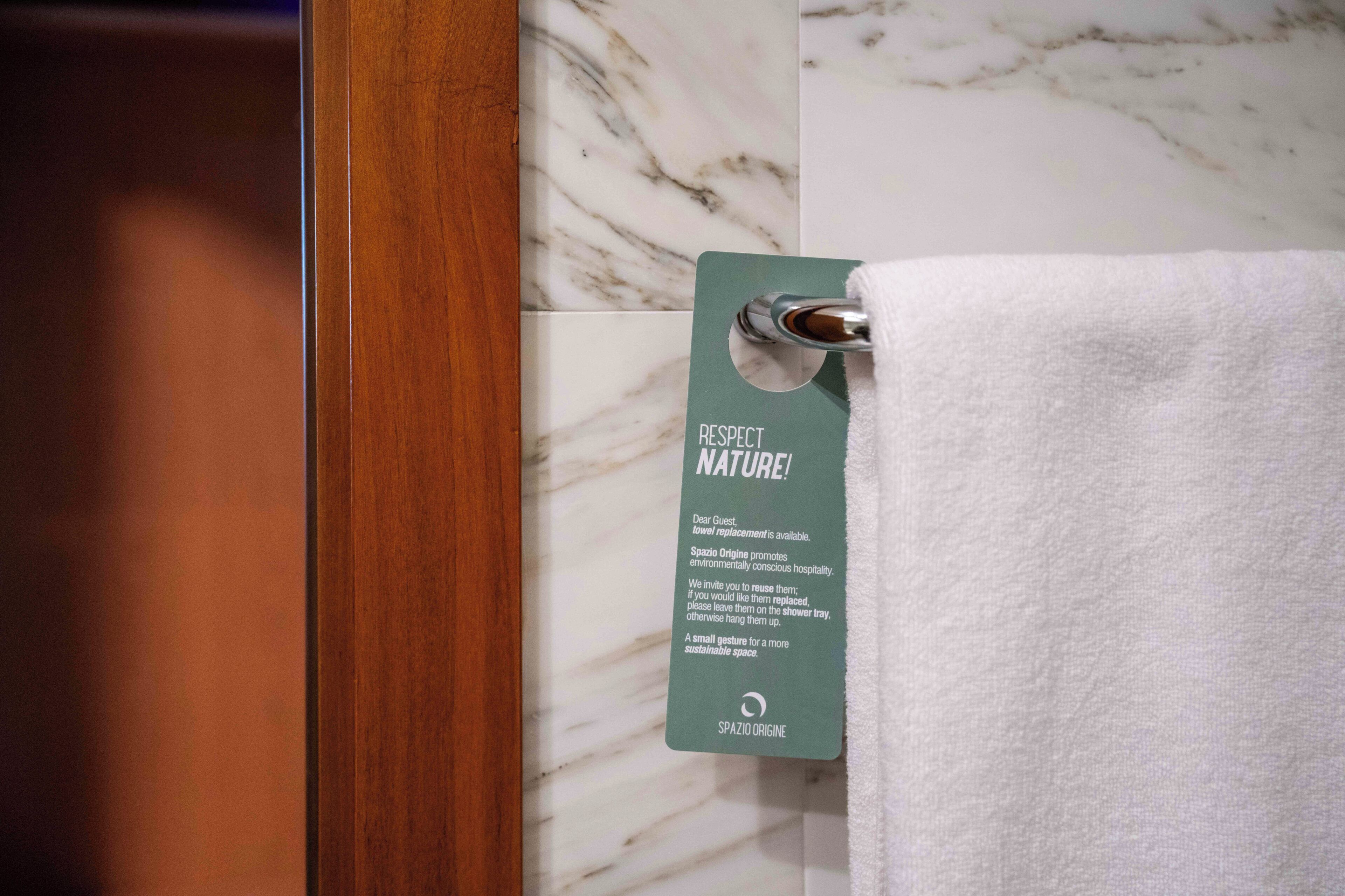Une salle de bain avec un mur en marbre, une porte en bois et une serviette suspendue à un crochet, ornée d’une étiquette sur laquelle on peut lire : « RESPECTONS LA NATURE ! »