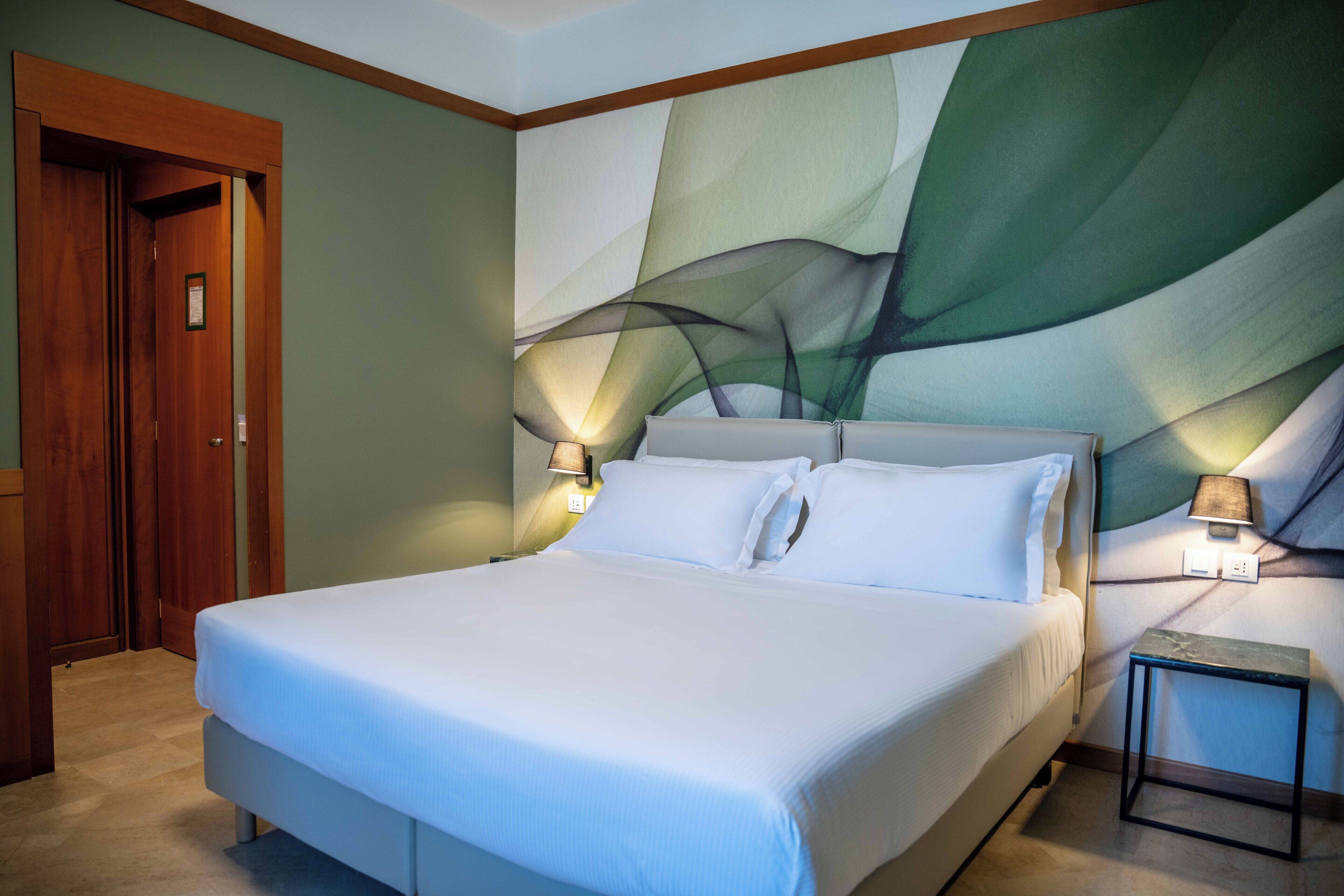 Quarto de hotel com uma cama grande, duas luminárias de cabeceira, uma porta de madeira e uma parede com arte abstrata.