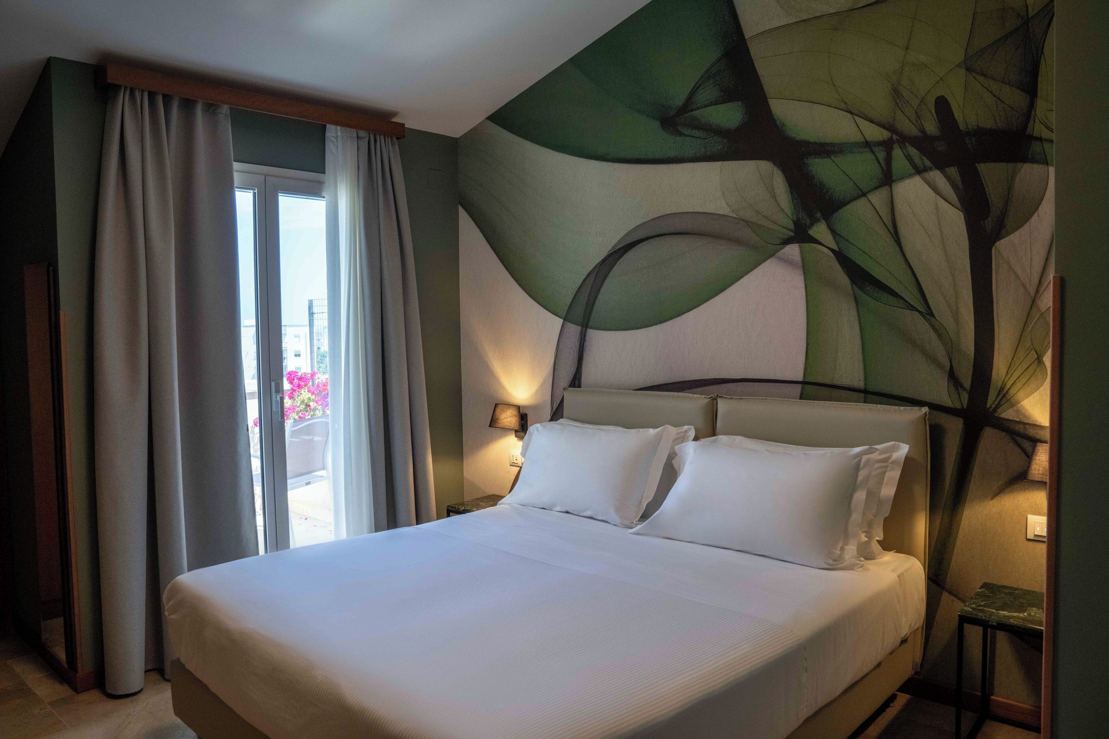 A hotel room with a bed, curtains, a nightstand with a lamp, and a wall with abstract art.