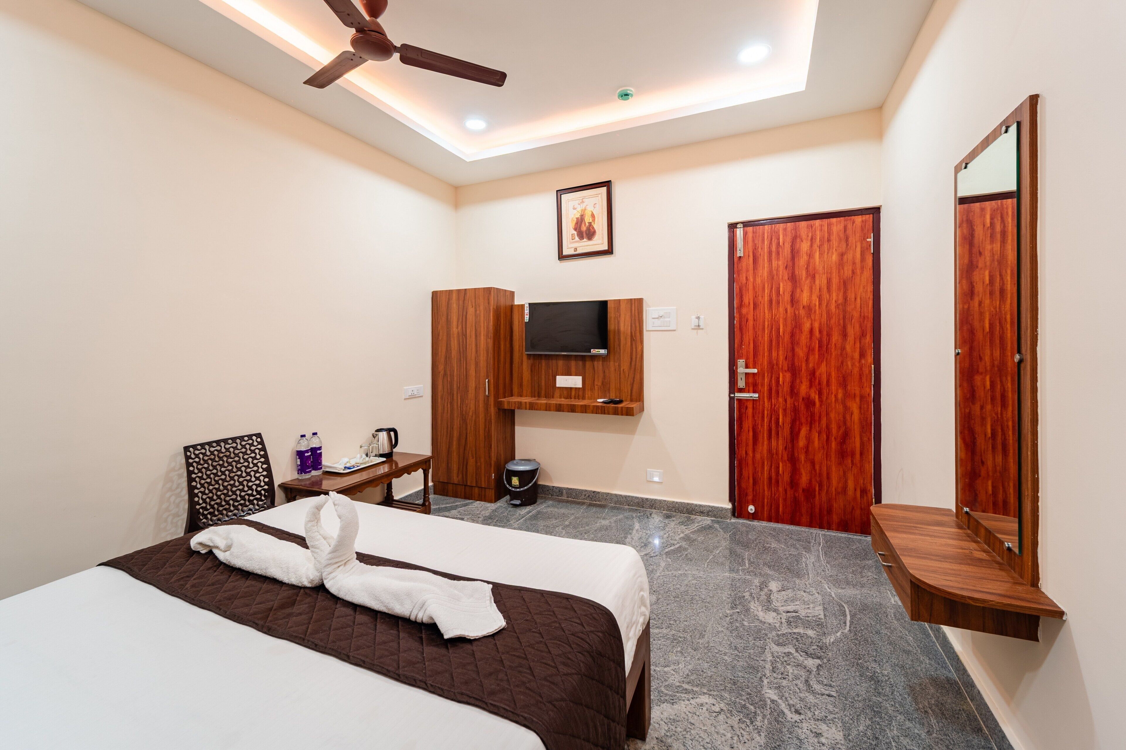 A hotel room with a bed, a ceiling fan, a mounted TV, wooden wardrobes, a small table with a chair, and a mirror.