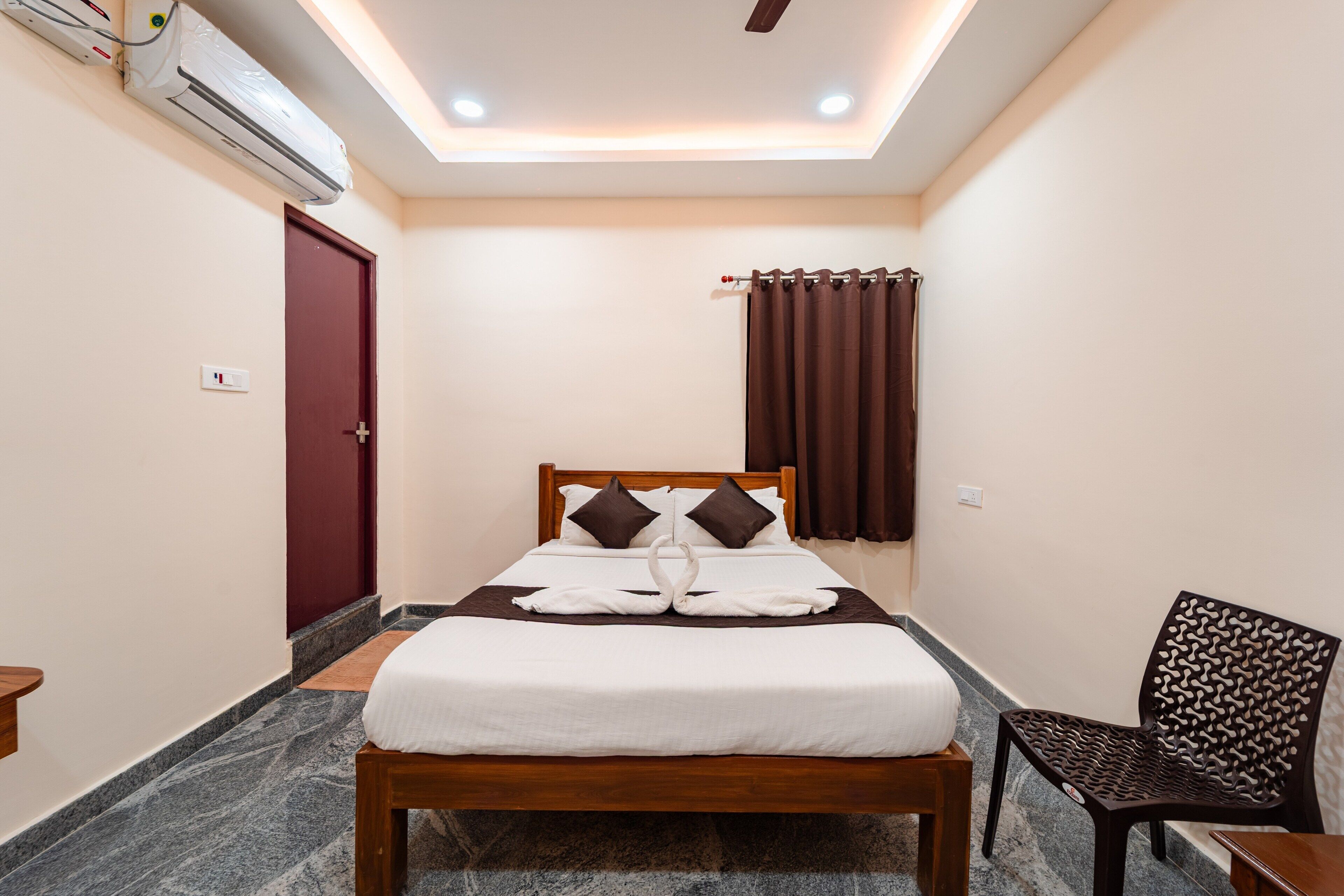 A hotel room with a bed, a wooden headboard, a brown curtain, a patterned chair, and an air conditioning unit.
