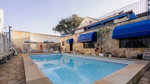 En swimmingpool med stenvægge og blå markiser, omgivet af udendørs siddepladser og en sti af sten.