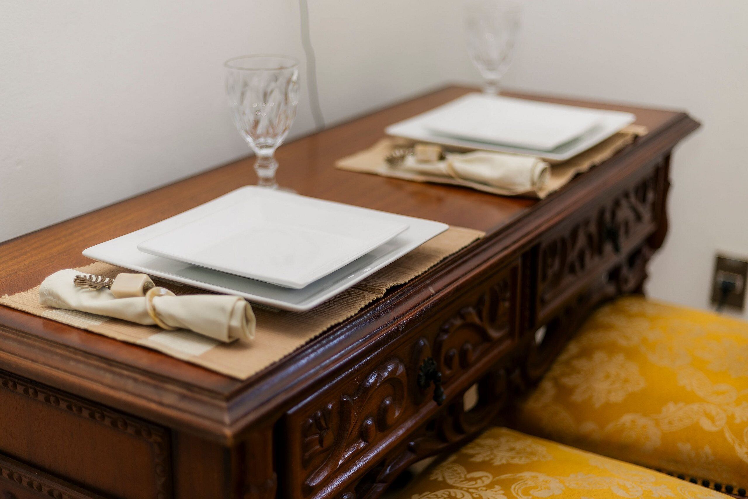 Une table à manger avec des assiettes, des couverts et des verres, placée sur une commode en bois surmontée d’un coussin jaune.