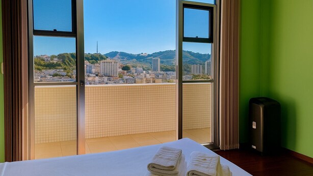 A hotel room with a bed, a view of a cityscape through large windows, and a suitcase. - Prime Center View - Beautiful view. Do it all on foot! (São Lourenço)