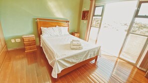 Een slaapkamer met een houten bed, nachtkastje, een dressoir en een groot raam met uitzicht.