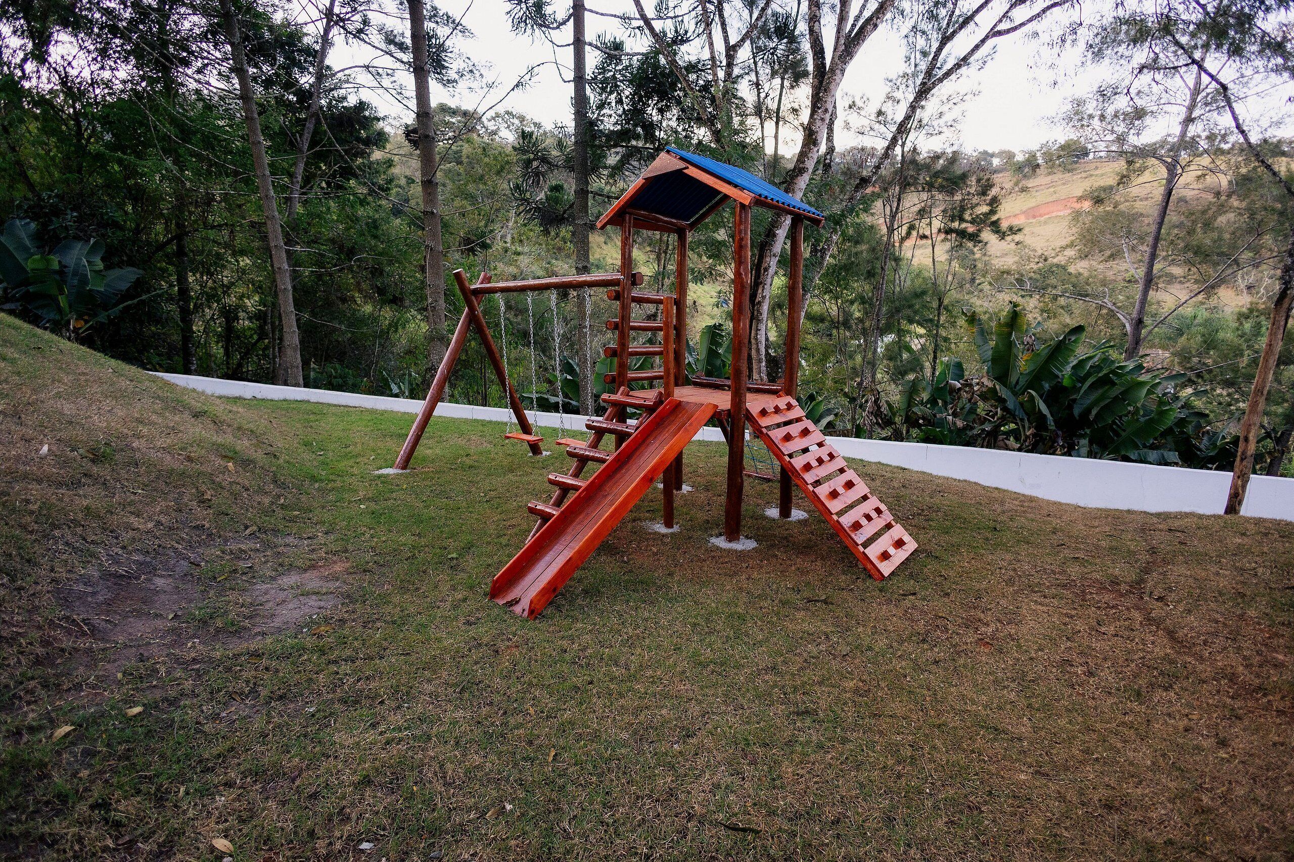 Um parquinho com escorrega e balanços, situado numa área gramada com árvores ao fundo.