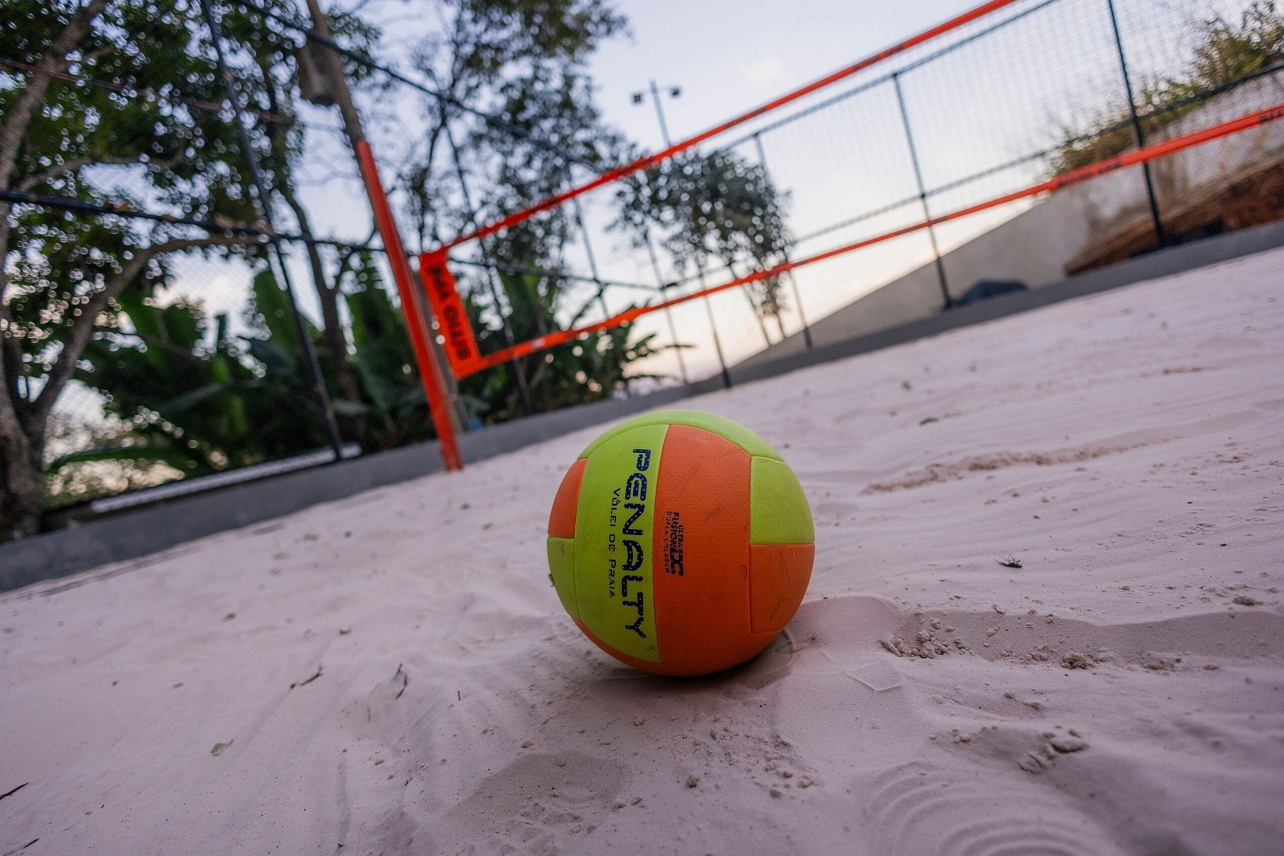Una pallavolo su una superficie sabbiosa, con una rete e una recinzione sullo sfondo.