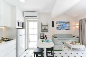 Eine moderne Küche mit weißen Schränken, einer eingebauten Mikrowelle und einem Kühlschrank. Es gibt einen Essbereich mit einem weißen Tisch und Stühlen, einer Couch und einem Couchtisch. Der Raum hat einen gefliesten Boden und ein an der Wand befestigtes Regal mit Pflanzen.
