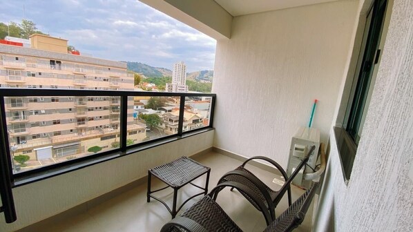 Un balcón con vistas a edificios, una pequeña mesa, dos sillas de mimbre y una ventana con marco de metal.