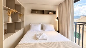 A hotel room with a bed, bedside tables, a nightstand, a lamp, a window with curtains, and a view of buildings outside.