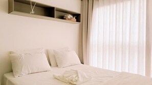 Quarto de hotel com cama, almofadas brancas, toalha branca dobrada, prateleira com livros e cortinas translúcidas.
