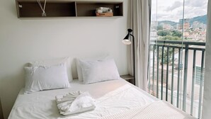 Ein Bett mit weißer Bettwäsche, ein Nachttisch mit Lampe, ein Bücherregal und Blick auf eine Stadtlandschaft durch einen Balkon.