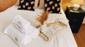 Una cama bien hecha con toallas blancas enrolladas, un jarrón decorativo con flores secas y una bandeja con un letrero.