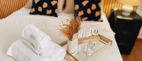 Een net opgemaakt bed met opgerolde witte handdoeken, een decoratieve vaas met gedroogde bloemen en een dienblad met een bordje.