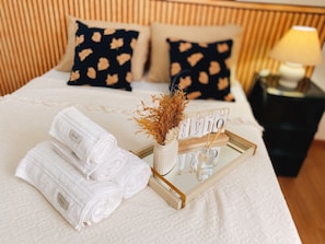 Una cama bien hecha con toallas blancas enrolladas, un jarrón decorativo con flores secas y una bandeja con un letrero.