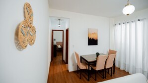 Una habitación con piso de madera, una mesa de comedor con sillas, una cama, una lámpara colgante y un cuadro en la pared.