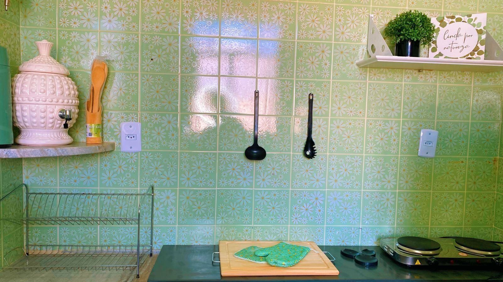 A kitchen with a green tiled backsplash, a wooden cutting board, a sink, and a shelf with kitchen utensils.