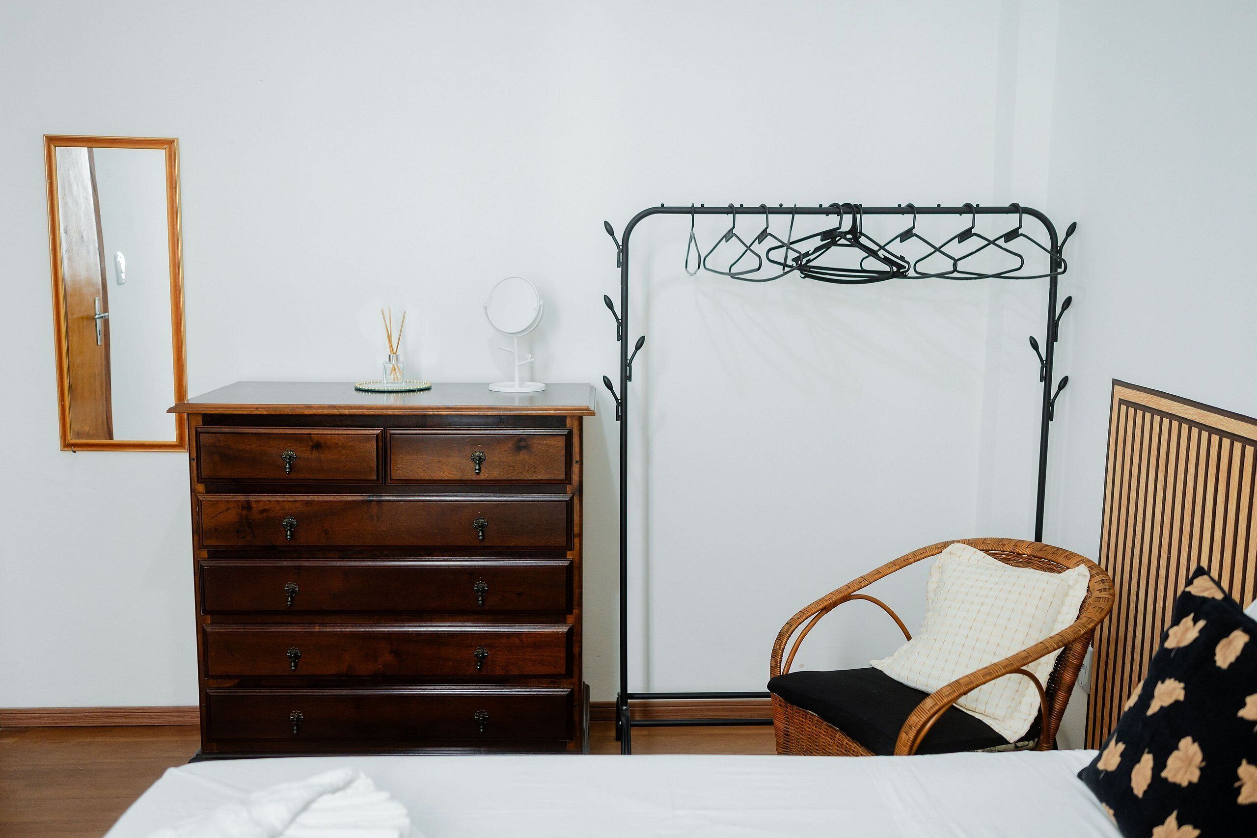Un dormitorio con cómoda de madera, perchero, silla de mimbre y cama con ropa blanca.