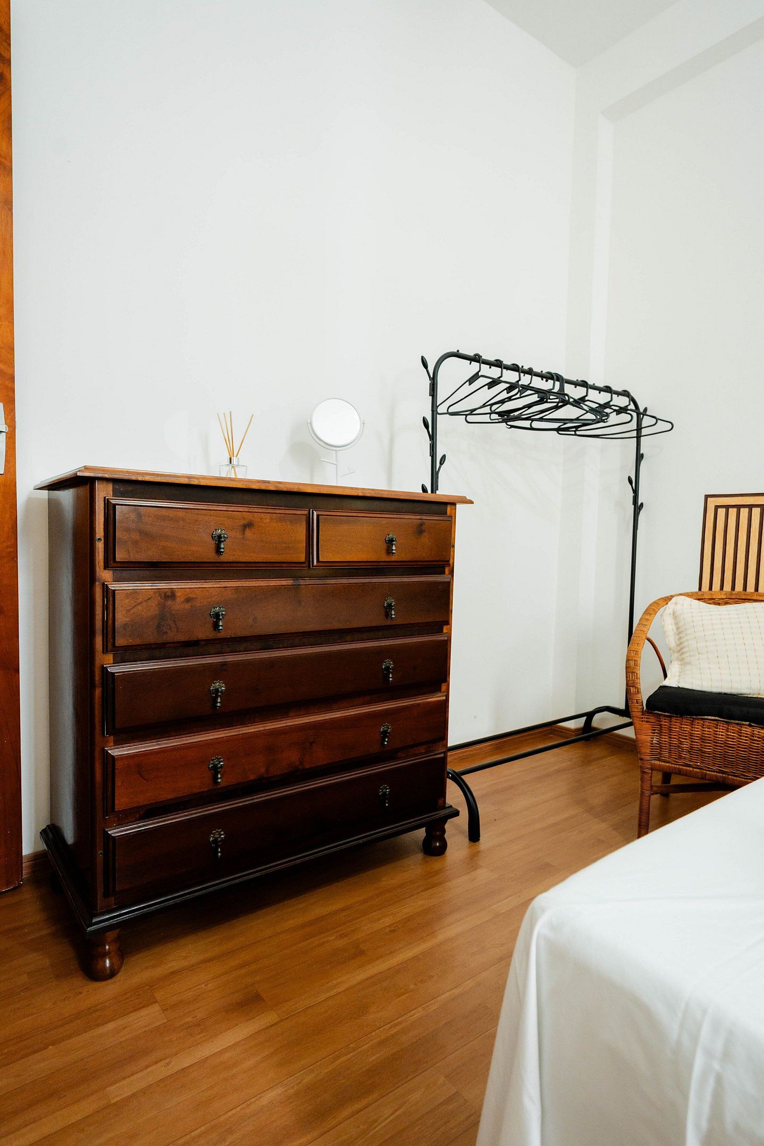Un aparador de madera con cajones, una silla de mimbre y una cama con sábana blanca.