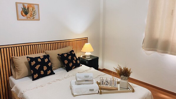 Una cama bien hecha con un cabecero a rayas, dos almohadas con estampado floral, una lámpara de noche, una bandeja con toallas y un calendario, y una obra de arte enmarcada en la pared.