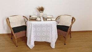 Una mesa de comedor con mantel blanco, dos sillas de mimbre con cojines negros y un florero con plantas secas sobre la mesa.