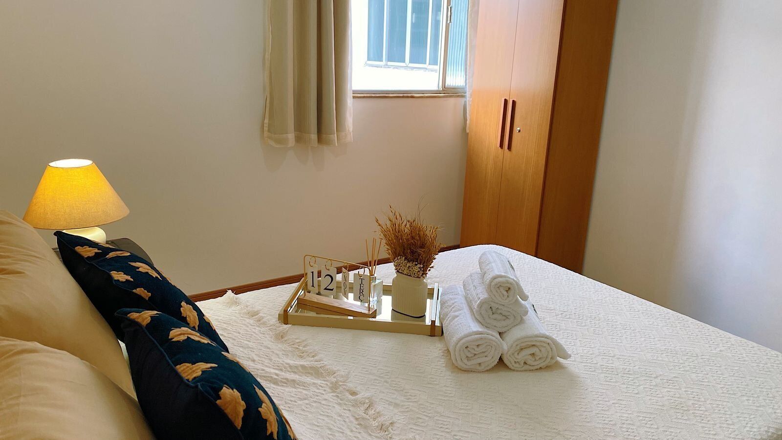 Una cama bien hecha con toallas enrolladas, una mesita de noche con lámpara, una bandeja con un calendario y objetos decorativos, y una ventana con cortinas.
