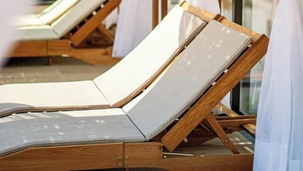 Una fila de sillas de sol con estructura de madera y cojines blancos, dispuestas en línea con cortinas blancas translúcidas.