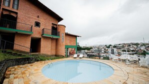 A swimming pool with a stone deck, a brick building with balconies, and a view of a town in the background. - Spacious Colonial House with Pool and Incredible View (São Lourenço)