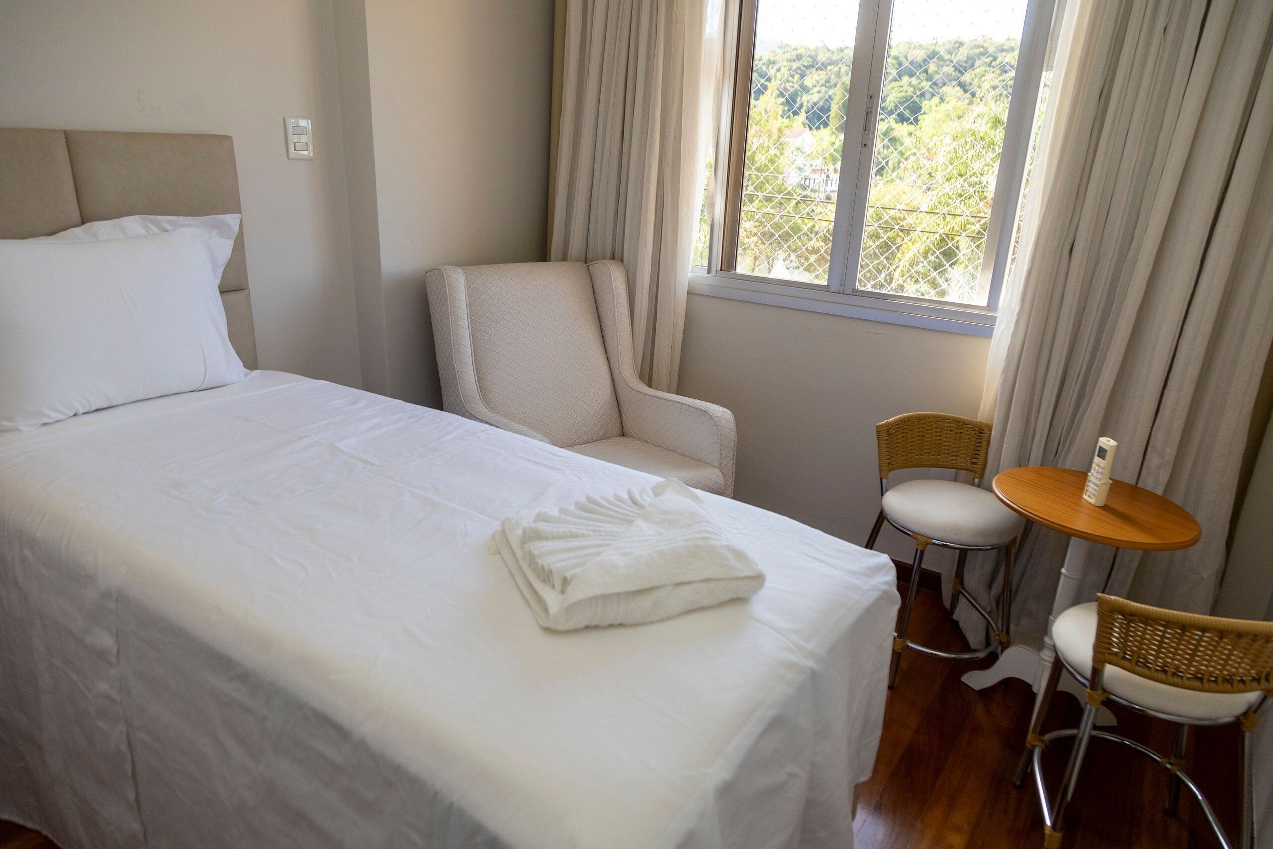 A hotel room with a bed, a chair, a small table, and a window with curtains.