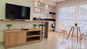 A modern kitchen with a flat-screen TV, wooden cabinetry, a dining area with a table and chairs, and a large window with sheer curtains.
