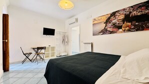 Ett sovrum med en säng, ett skrivbord, en stol, en tv och en målning som föreställer en kuststad.