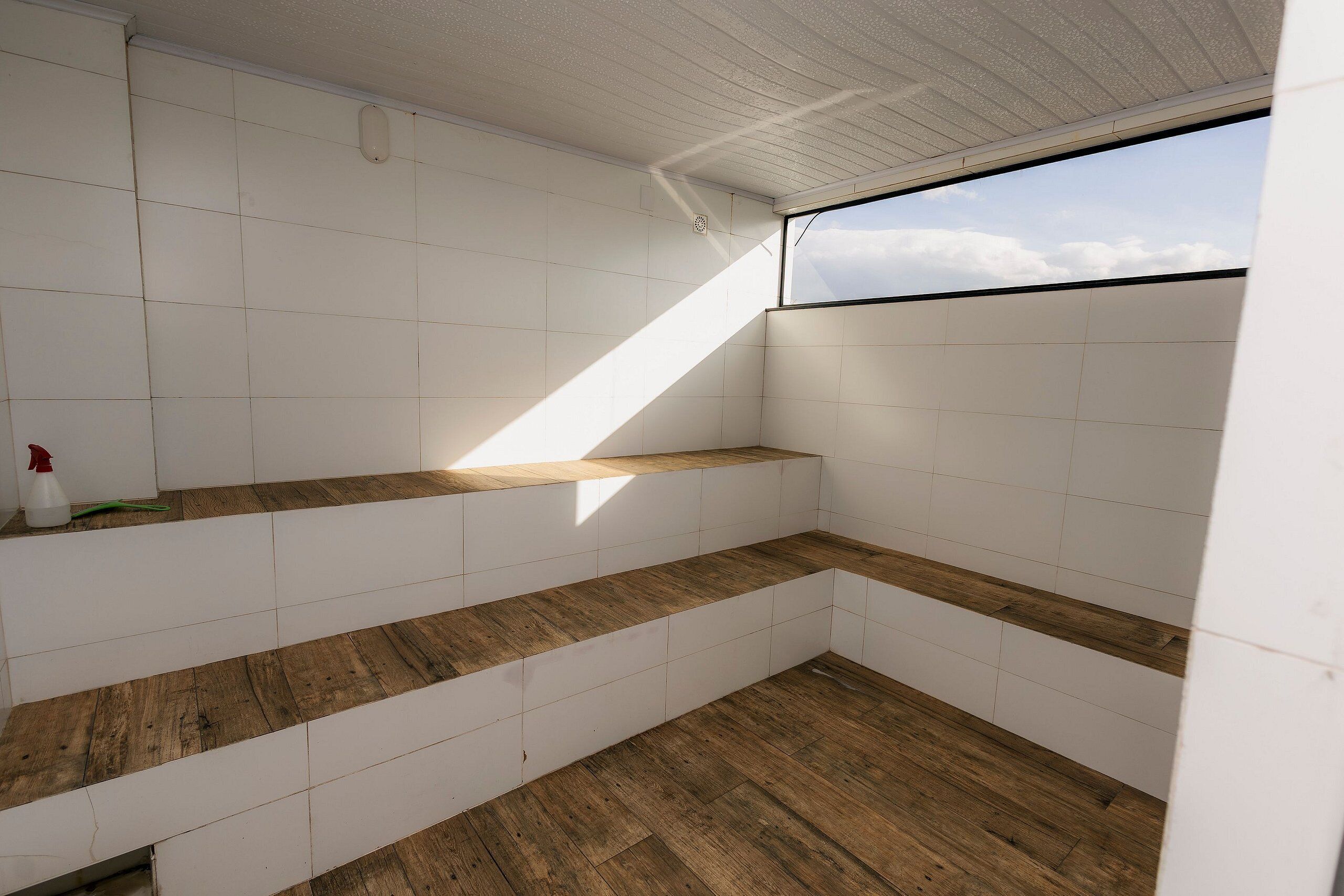 Esquina de una habitación con paredes de azulejos blancos, piso de madera y ventana con vista al cielo.