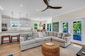 A modern living room with a sectional sofa, a round woven ottoman, and a dining area with a view of the outdoors. - Coastal Livin' by Duncan Real Estate (Holmes Beach)