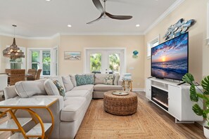 A living room with a sectional sofa, a TV, a ceiling fan, and a dining area with wicker furniture. - Coastal Livin' by Duncan Real Estate (Holmes Beach)