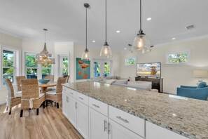 A spacious kitchen with a large island, dining area with wicker chairs, and a living area with a blue sofa. - Coastal Livin' by Duncan Real Estate (Holmes Beach)