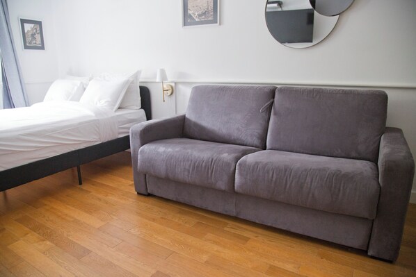 A room with a bed, a grey sofa, a round mirror, and a wall-mounted lamp. - Nice renovated AC studio in Saint Germain (Paris)