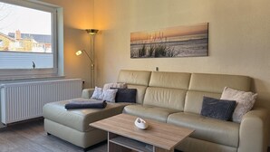 A living room with a sofa, a coffee table, a floor lamp, and a painting on the wall. - Haus Nordseekrabbe Apartment 11 46m2 Terrace Wheelchair accessible Category 2 (Neuharlingersiel)