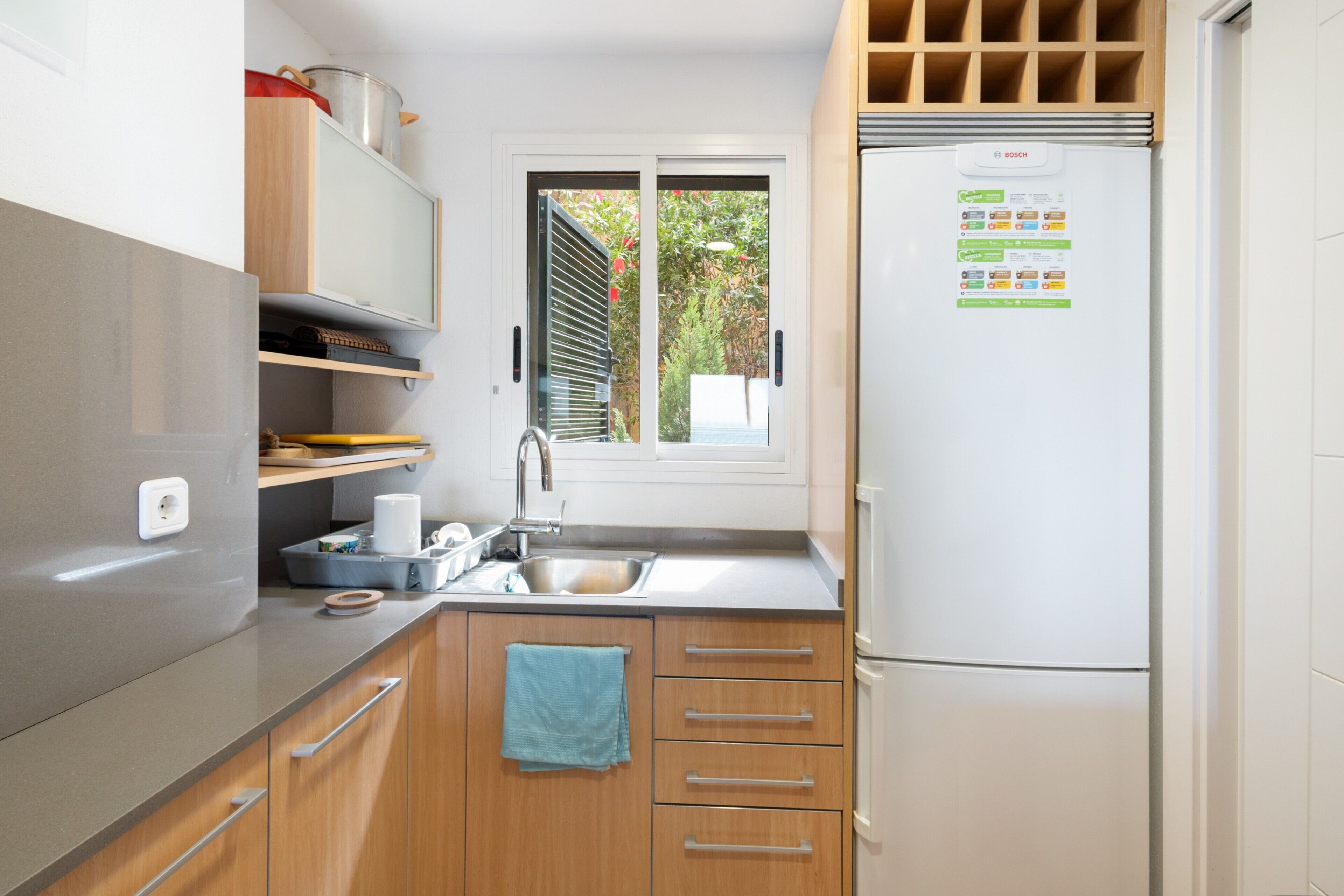 Een compacte keuken met een Bosch koelkast, een spoelbak, houten kastjes en een raam met zonwering.