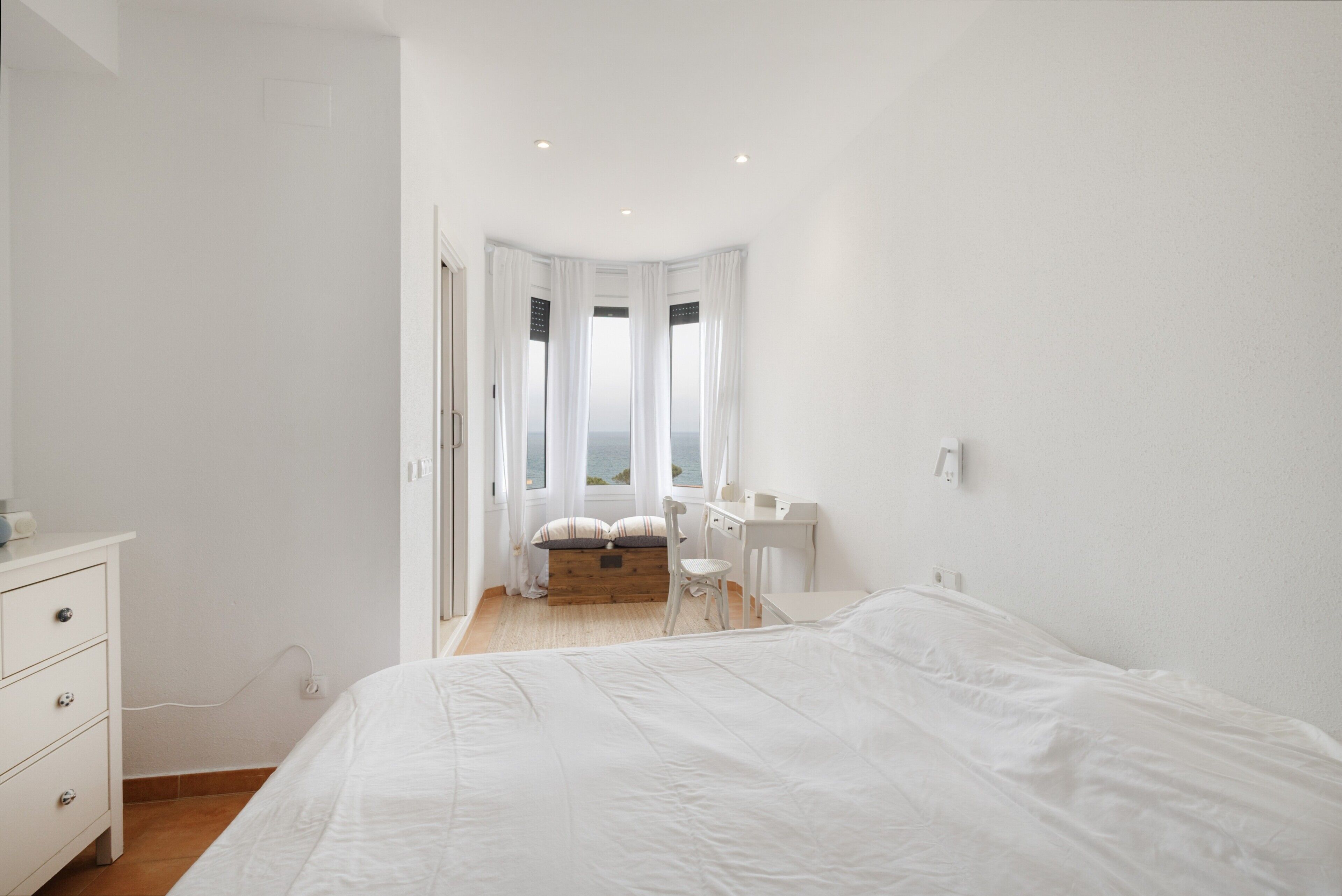 A bedroom with a bed, a desk, a chair, and a view of the sea through a window.