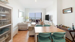 A room with a dining table, chairs, a TV, a bookshelf, and a view of the sea. - Apartment 'Sa Calma 2 - 1D' with Sea View, Wi-Fi and Air Conditioning (Begur)