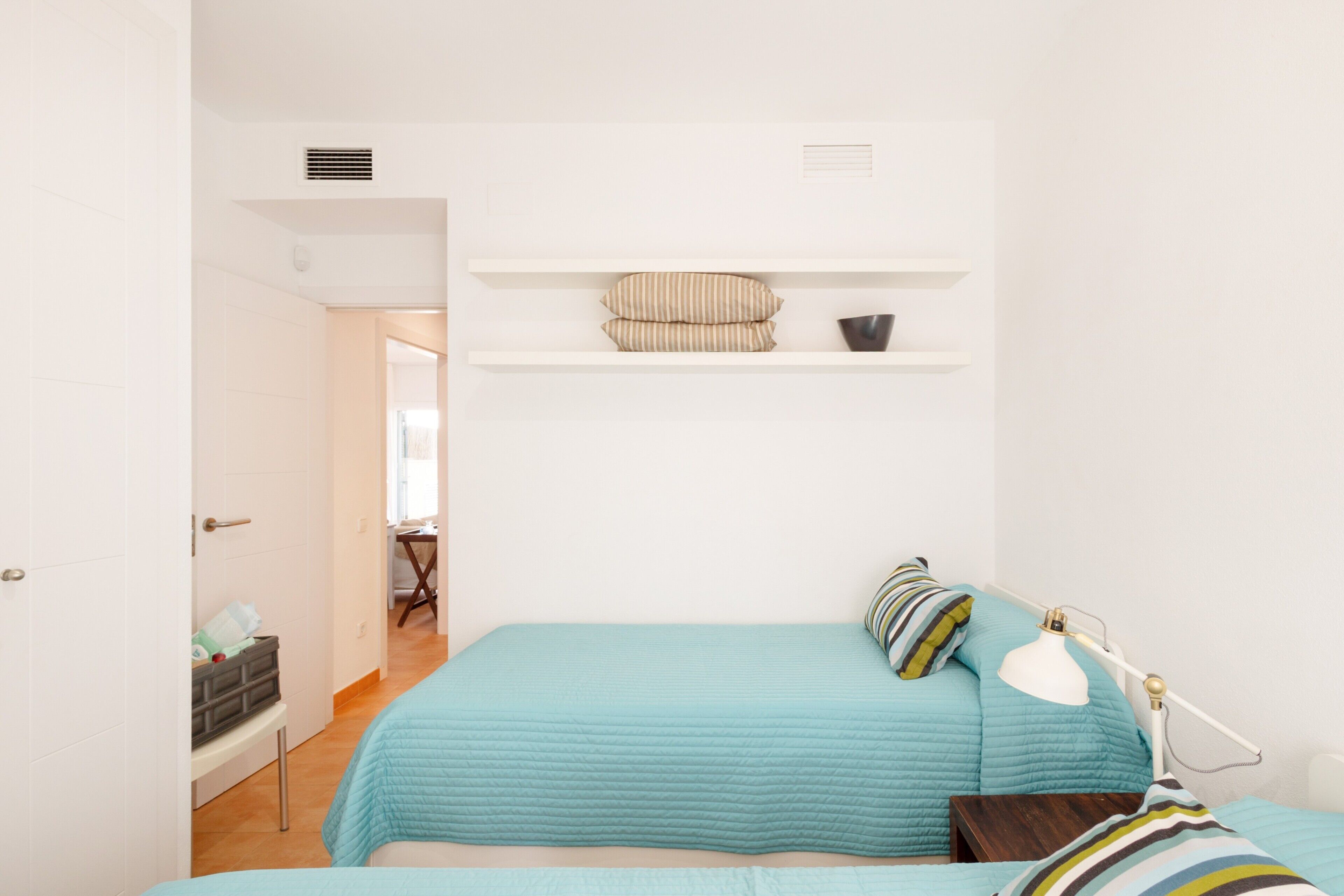 A bedroom with a bed, a bedside table, a lamp, and a shelf with a pillow and a bowl.