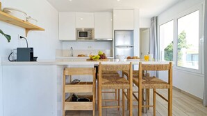 Una cocina moderna con armarios blancos, un refrigerador de acero inoxidable, un microondas y un frutero en la encimera. Hay tres sillas de mimbre alrededor de una pequeña isla con mesa.