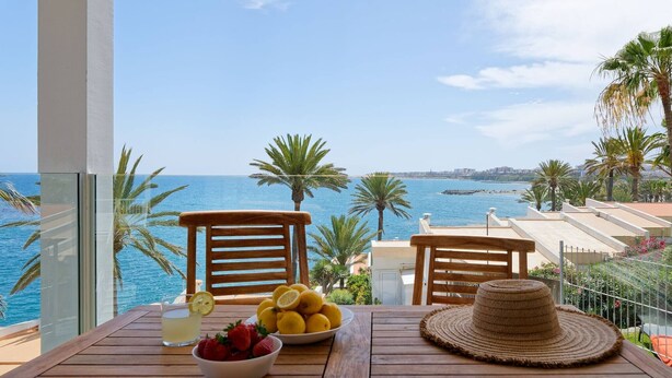 Una mesa de madera con un bol de fresas, un vaso de bebida y un plato de limones, con vistas a la costa, palmeras y un cielo despejado.