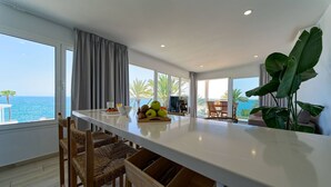 Una cocina moderna con una isla central, comedor con sillas, ventanas amplias con vistas al océano y una planta.