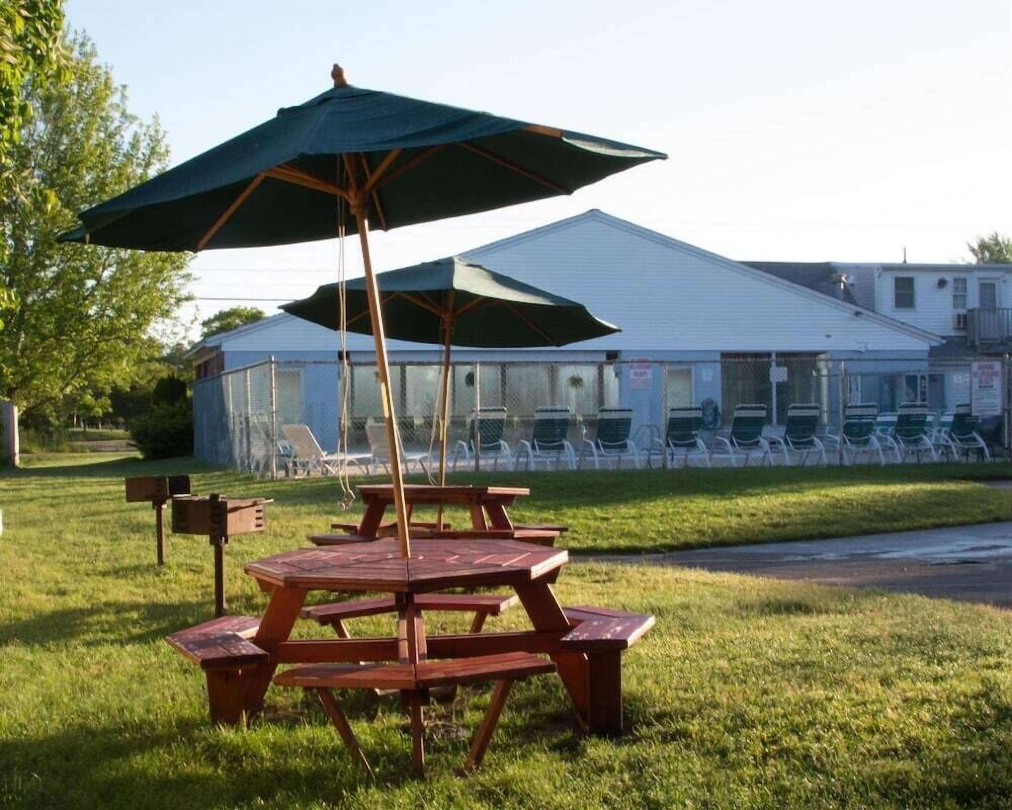 A picnic table with a green umbrella, a grassy area, and a building with a white roof in the background.