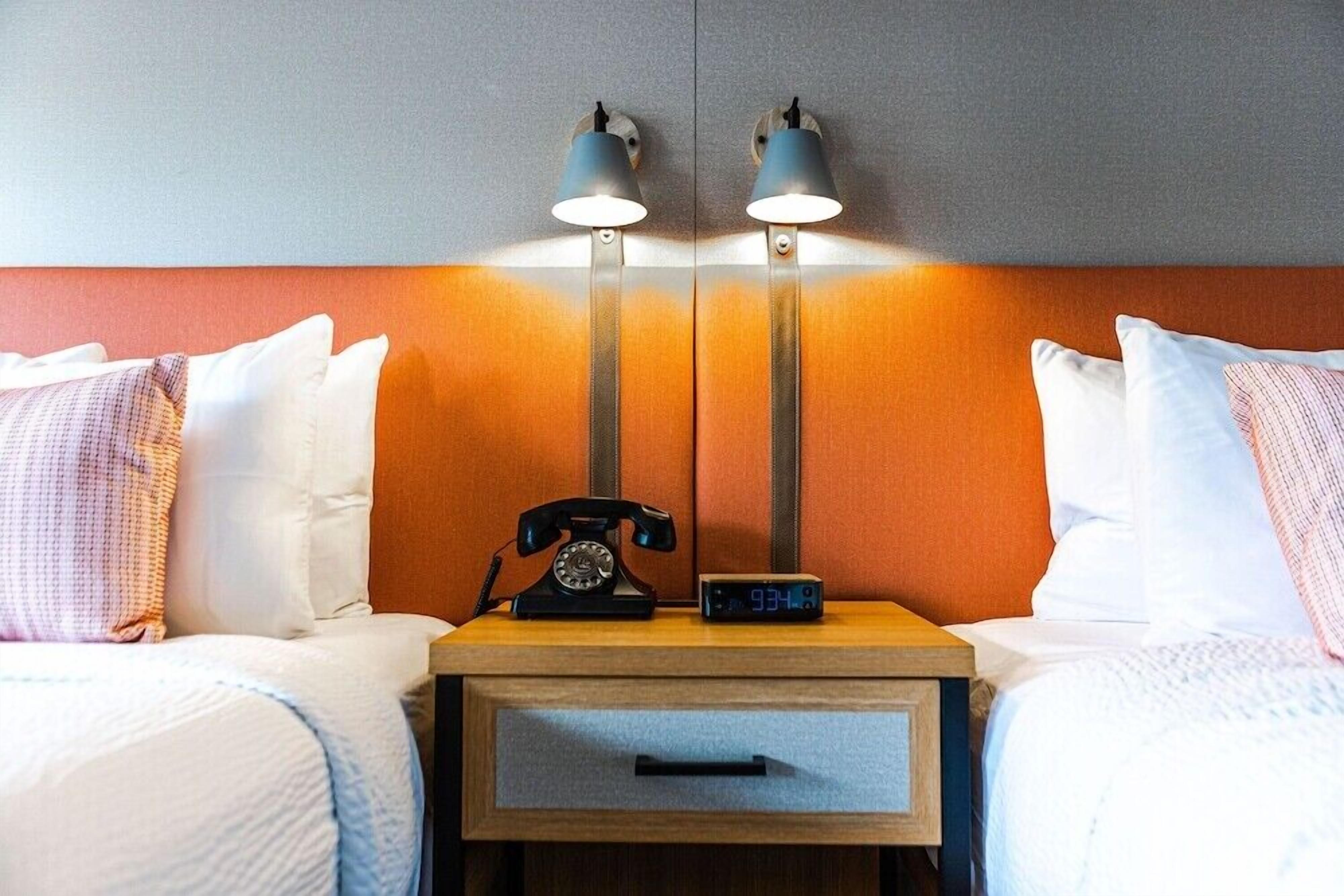 A hotel room with two beds, a nightstand with a telephone and a digital clock, and wall-mounted lamps.