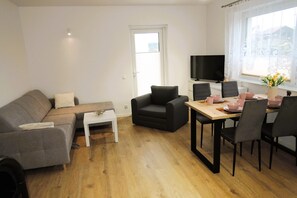 Ein Wohnzimmer mit Sofa, einem kleinen Tisch, einem Esstisch mit Stühlen, einem Fernseher und einem Fenster mit Vorhängen.